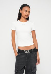 Fehér, rövid ujjú, testhezálló póló kerek nyakkal és ezüst logó részlettel a mellkason, sötétszürke cargo nadrággal és fekete övvel párosítva.