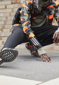 Abbigliamento sportivo da esterno che presenta un giubbotto con motivo geometrico nero, arancione e verde, pantaloni neri e scarpe da ginnastica nere e arancioni con texture.