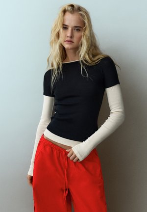 Un mannequin blond pose devant un mur clair, vêtu d’un haut noir à manches courtes par-dessus un vêtement blanc à manches longues et d’un pantalon rouge vif.