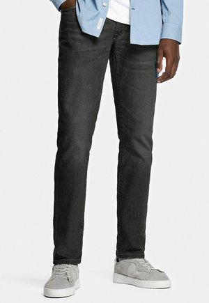 Man draagt nauwsluitende zwarte spijkerbroek, lichtgrijze sneakers, wit overhemd en lichtblauw jasje, staat met één hand in de zak.