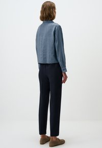 Blauwe textuurjas met lange mouwen en gecolloceerde kraag, gecombineerd met donkere broek en platte schoenen. De achterzijde toont het patroon van de jas.