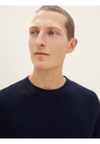Marineblauer Strickpullover mit strukturiertem Muster, rundem Halsausschnitt und gerippten Ärmeln. Nahaufnahme der Stoff- und KragenDetails.