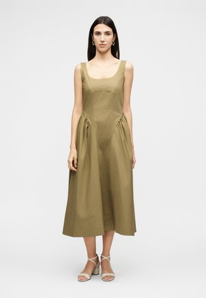 Femme aux cheveux raides et foncés portant une robe midi sans manches vert olive et des sandales à talons beiges, debout devant un fond clair uni.