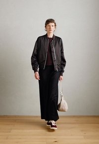 Black satin bomber jacket over a burgundy top, paired with wide black trousers and burgundy sneakers. Light gray bag with a strap.