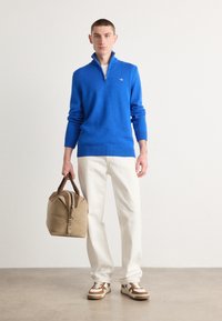 Blauer gestrickter Pullover mit hohem Kragen, kombiniert mit weißen, locker sitzenden Hosen und Sneakers mit braunen Akzenten. Hält eine beige Reisetasche.