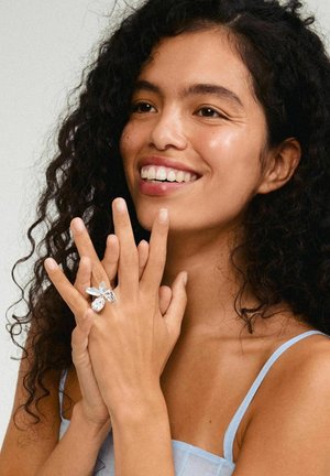 Anello in argento con un design dettagliato di farfalla e foglie testurizzate, indossato su una mano con unghie naturali, su uno sfondo neutro.