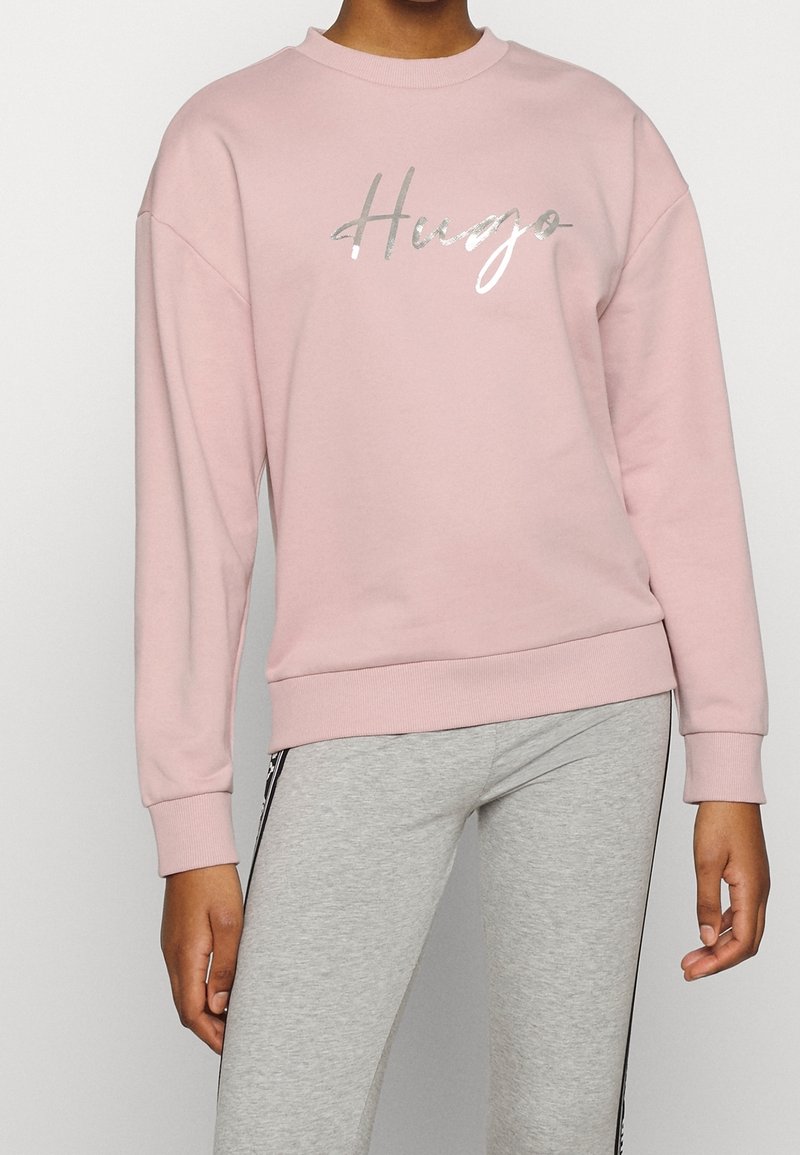 Rosa Sweatshirt mit Rundhalsausschnitt, ausgestattet mit einem silbernen Logo "Hugo" in Schreibschrift. Hergestellt aus weichem Material mit lässiger Passform und gerippten Bündchen.