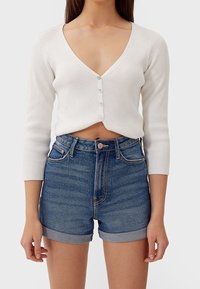 White ribbed cropped cardigan with long sleeves and a deep V-neck, buttoned at the front. Paired with high-waisted blue denim shorts.