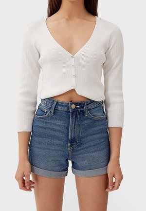 Weißes geripptes cropped Cardigan mit langen Ärmeln und tiefem V-Ausschnitt, vorne geknöpft. Kombiniert mit hoch taillierten blauen Denim-Shorts.