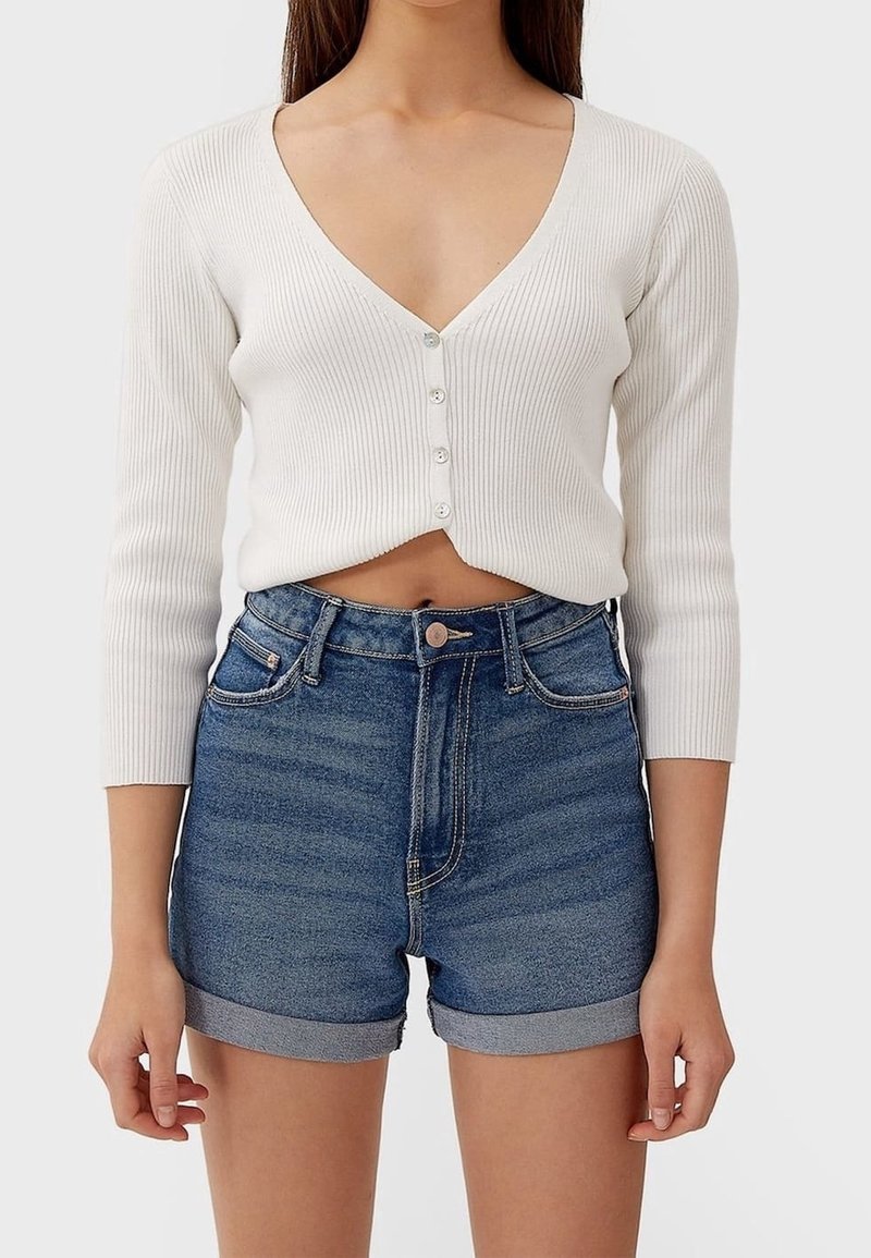 White ribbed cropped cardigan with long sleeves and a deep V-neck, buttoned at the front. Paired with high-waisted blue denim shorts.