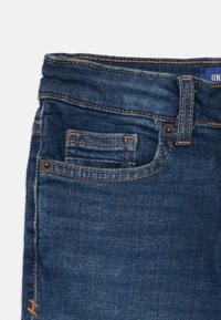 Dunkelblaue Denim-Jeans mit einer glatten Textur, die orangefarbene Nähte, eine kleine Fronttasche und einen Knopfverschluss an der Taille aufweisen.