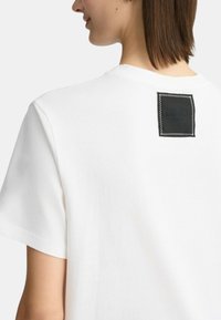 Maglietta in cotone bianco con collo rotondo e maniche corte. Etichetta in pelle nera ben visibile sulla parte posteriore con il logo "LIU•JO" in rilievo. Texture liscia.