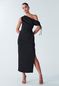 Juoda vienspetė suknelė iš lygaus audinio, su šoniniu skeltuku ir rauktu detalės elementu, derinama su auksinėmis dirželiais papuoštomis sandalais.