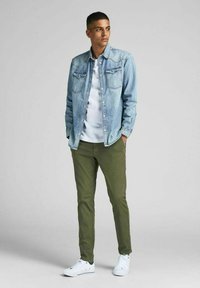 Giacca in denim azzurro chiaro con doppie tasche sul petto, abbinata a una t-shirt bianca e chinos verdi; completato con sneakers bianche.