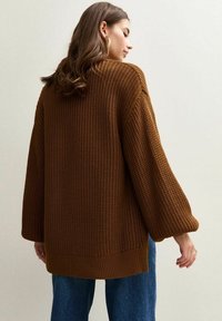 Oversized brown knit sweater with textured fabric, dropped shoulders, and wide, long sleeves. Ribbed hem adds detail. Worn with blue jeans.
