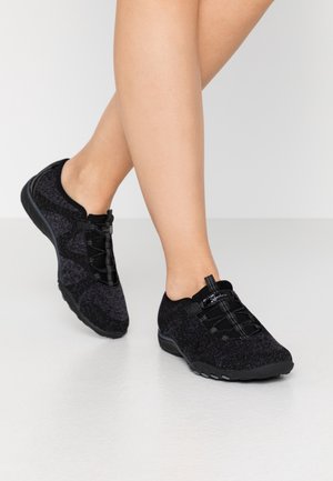 Jambes chaussées de baskets noires sans lacets avec des lacets élastiques, posées sur un sol blanc, un pied légèrement levé en plein pas.