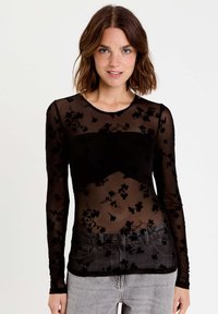 Haut sheer noir à manches longues avec broderie en velours floral, col rond et design ajusté, associé à un jean gris taille haute.