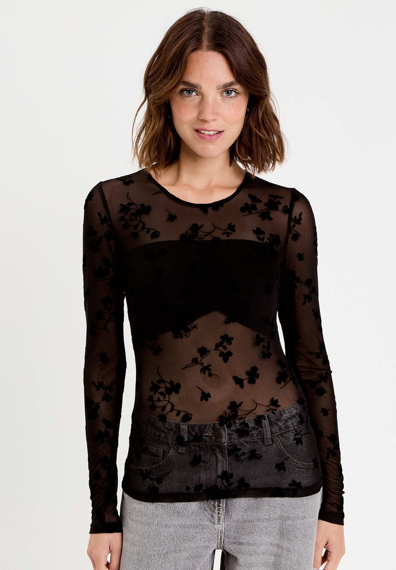 Haut sheer noir à manches longues avec broderie en velours floral, col rond et design ajusté, associé à un jean gris taille haute.