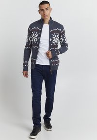 Graue Strickjacke mit hohem Kragen, weißem und braunem Schneeflockenmuster, gerippten Manschetten und Reißverschluss, getragen über einem weißen Hemd und blauen Jeans.