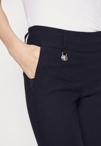 Pantaloni blu navy con una texture liscia, dotati di una vita con cordoncino, tasche laterali e un dettaglio in metallo in vita.