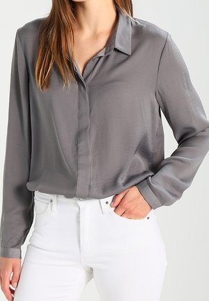 Frau trägt ein graues Langarmhemd mit Knöpfen, das in eine weiß hochtaillierte Hose gesteckt ist, eine Hand in der Tasche.