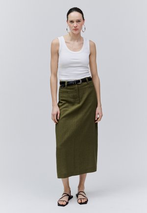 Mujer con camiseta blanca sin mangas, falda midi de talle alto color verde oliva con cinturón negro, grandes pendientes de aro y sandalias negras de tiras, de pie en posición neutral.