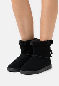 Stivaletti neri alla caviglia in morbido suede, con un bordo in pelliccia sintetica e un fiocco decorativo sul retro, dotati di una suola in gomma testurizzata.