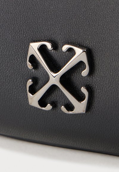 Emblème métallique avec quatre pointes de flèches symétriques dirigées vers l'extérieur sur une surface noire texturée, probablement un logo ou un détail décoratif.