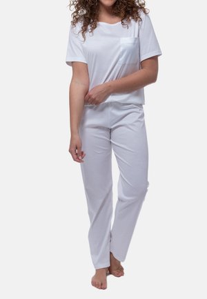 Vrouw in wit shirt met korte mouwen en zak en witte broek, op blote voeten staand tegen een effen achtergrond.