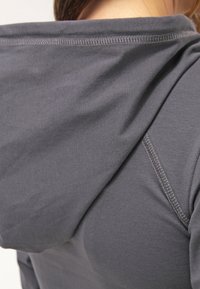 Gros plan d’une personne portant un sweat à capuche gris, montrant les coutures détaillées de la capuche et de la couture de l’épaule.