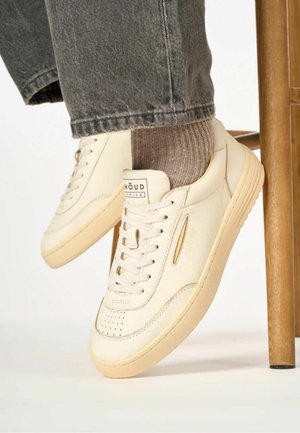 Sneaker in pelle crema con punta perforata, dettagli beige e logo dorato. Indossate con jeans grigi e calzini a coste marroni, posizionate vicino a una sedia di legno.