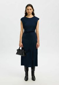Femme portant une robe midi sans manches couleur marine, des bottes noires jusqu'aux genoux, tenant un petit sac à main noir devant un fond blanc uni.