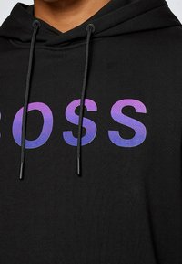 Černá mikina s kapucí s velkým gradientním fialovým nápisem "BOSS." Obsahuje šňůrky s černými konci a měkkou, strukturovanou tkaninu.