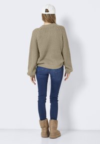 Beige stickad tröja med avslappnad passform, rund halsringning och ribbade ärmslut, kombinerad med smala blå jeans och bruna stövlar. Vy från baksidan.