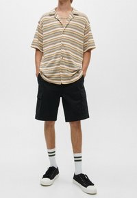 Gestreept T-shirt met korte mouwen in beige, bruin en marineblauw; gecombineerd met zwarte cargoshorts. Zwarte sneakers en witte sokken met groene strepen.