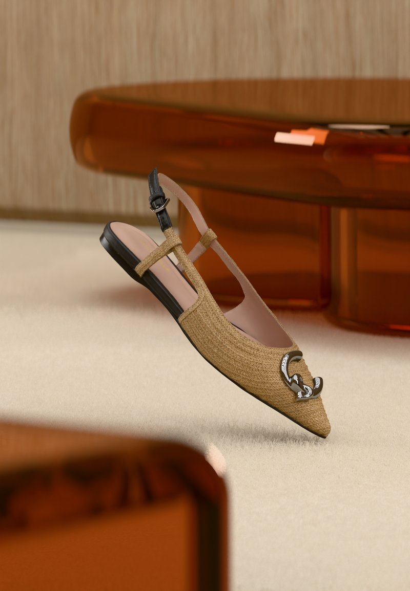 Μπεζ πλεκτό παπούτσι φλατ με λουράκι στο πίσω μέρος, μαύρη αγκράφα και ασημένιο διακοσμητικό έμβλημα στην μυτερή μύτη, εκτεθειμένο σε εσωτερικό χώρο πάνω σε εκρού μοκέτα.