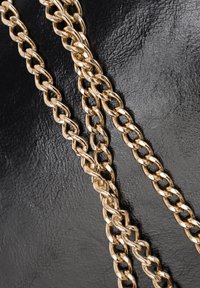 Złote ogniwa metalowego łańcucha mają polerowane wykończenie i zaprojektowane są jako zazębiające się elementy. Tłem jest czarna, teksturowana skóra.