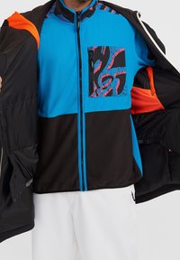 Homme portant une veste noire et bleue zippée avec une poche poitrine à motif, ouvrant une veste extérieure noire avec une doublure orange. Pantalon blanc visible en dessous.