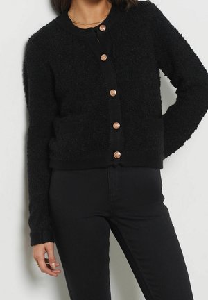 Vrouw draagt een zwarte zachte cardigan met gouden knopen, zwarte broek en een delicate gouden ketting, poseren met één hand op haar heup.