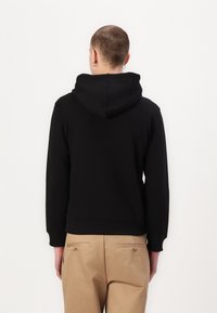Sweatshirt preto com capuz, de ajuste relaxado, com punhos e bainha canelados. Combinado com calças bege com bolsos traseiros e detalhes dourados.