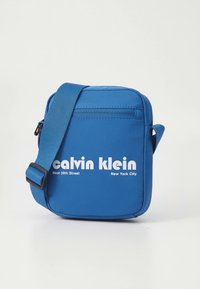 Blå Calvin Klein skulderveske med justerbar stropp, frontlomme med glidelås og hvit tekst som sier "West 39th Street New York City."