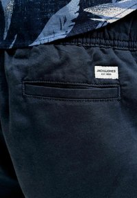 Κοντινή λήψη μαύρου casual παντελονιού με πίσω τσέπη με σχισμή και λευκή ετικέτα "JACK & JONES EST.1990" κοντά στη μέση.