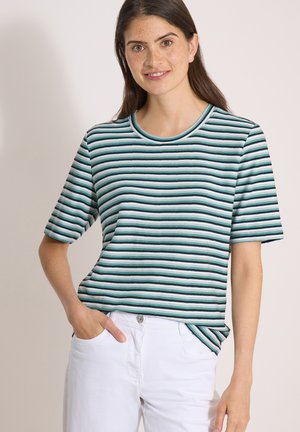 Jeune femme aux cheveux bruns portant un t-shirt rayé vert, noir et blanc et un pantalon blanc, debout avec une main dans la poche.