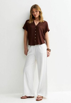 Vrouw draagt een bruin, kort mouwig, knoopblouse, witte broek met wijde pijpen, armbanden en bruine sandalen, staand tegen een witte muur.