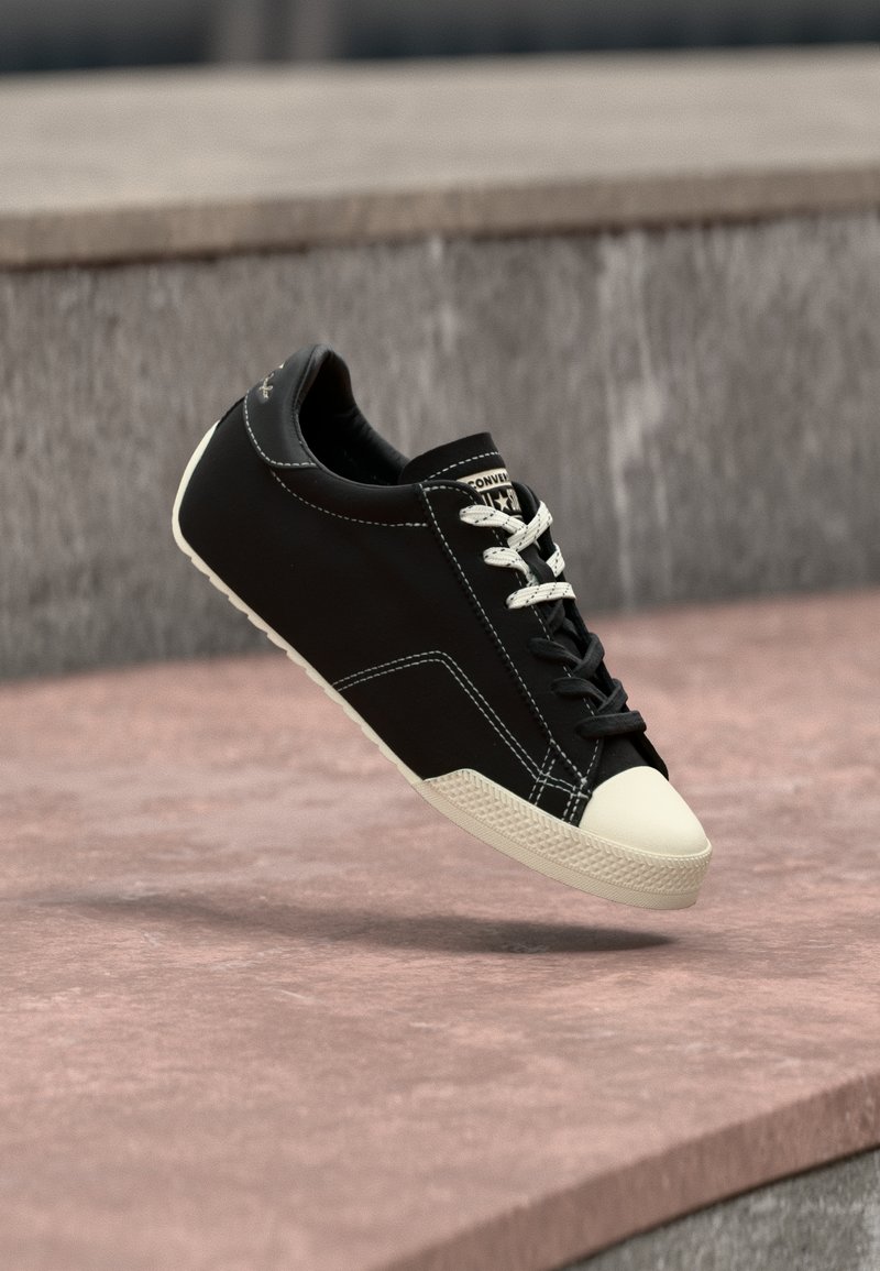 Μαύρο αθλητικό παπούτσι με λευκό δάχτυλο και κορδόνια φαίνεται να αιωρείται πάνω από μια ροζέ απόχρωση τσιμεντένια επιφάνεια με γκρι σκαλοπάτια στο φόντο.