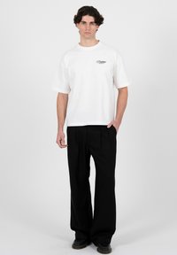 Mann mit dunklem, lockigem Haar trägt ein weißes, übergroßes T-Shirt mit Aufschrift und lockere schwarze Hose, steht vor einem schlichten grauen Hintergrund.