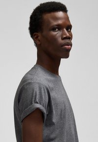 Nuori mies, jolla on lyhyet kiharat hiukset ja hänellä on harmaa t-paita, jonka hihat on kääritty ylös, katsoo sivulle yksinkertaisen vaalean taustan edessä.