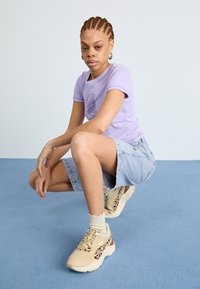 T-shirt lilás clara ajustada com estampado, calções de ganga até ao joelho e ténis bege com detalhes em padrão de leopardo. Meias brancas caneladas visíveis.