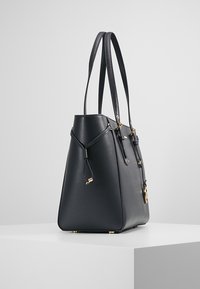 Musta tekstuuripintainen nahkalaukku, jossa on kaksi pitkää kahvaa, suppilomainen muoto, kultaiset metalliosat ja turvallinen suljentayksityiskohta.