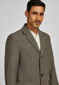 Man met een bruine gestructureerde blazer over een wit knoopschoenhemd, recht vooruit kijkend met een neutrale uitdrukking tegen een effen achtergrond.